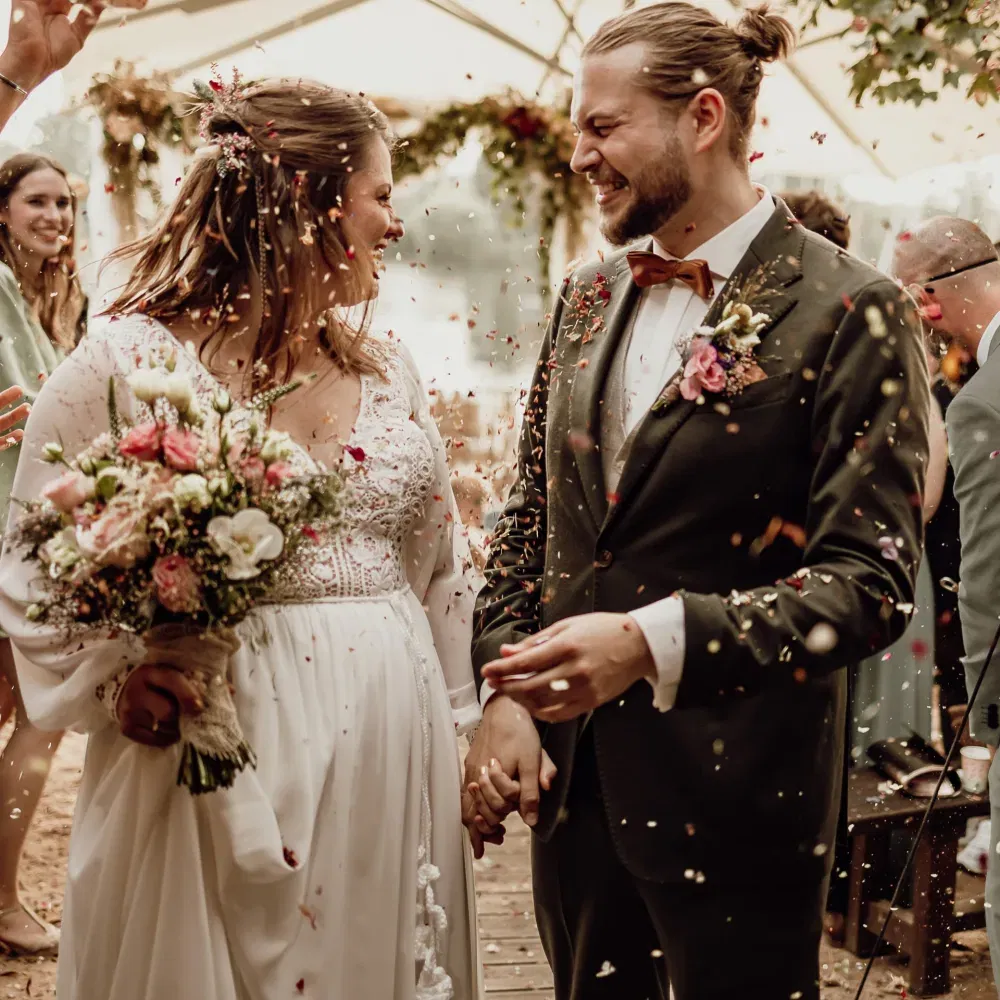 Frisch verheiratetes Paar geht lächelnd Hand in Hand durch ein Spalier, während Gäste farbige Blütenkonfetti über ihnen verstreuen.