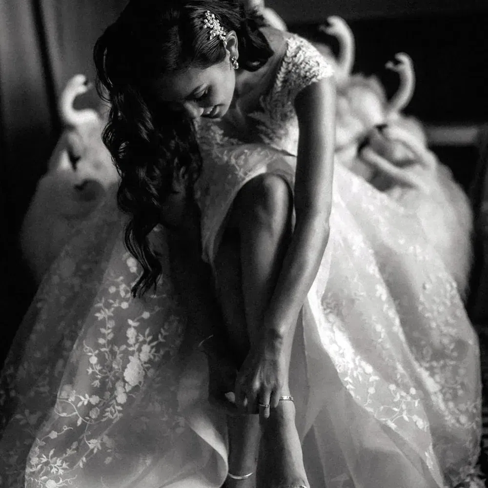 Schwarz-weiß-Aufnahme einer Braut, die sich im Sitzen ihre Sandalen schließt. Sie trägt ein besticktes, ausladendes Spitzen-Brautkleid und hat wellige Haare mit einem funkelnden Haaraccessoire zur Seite gesteckt.