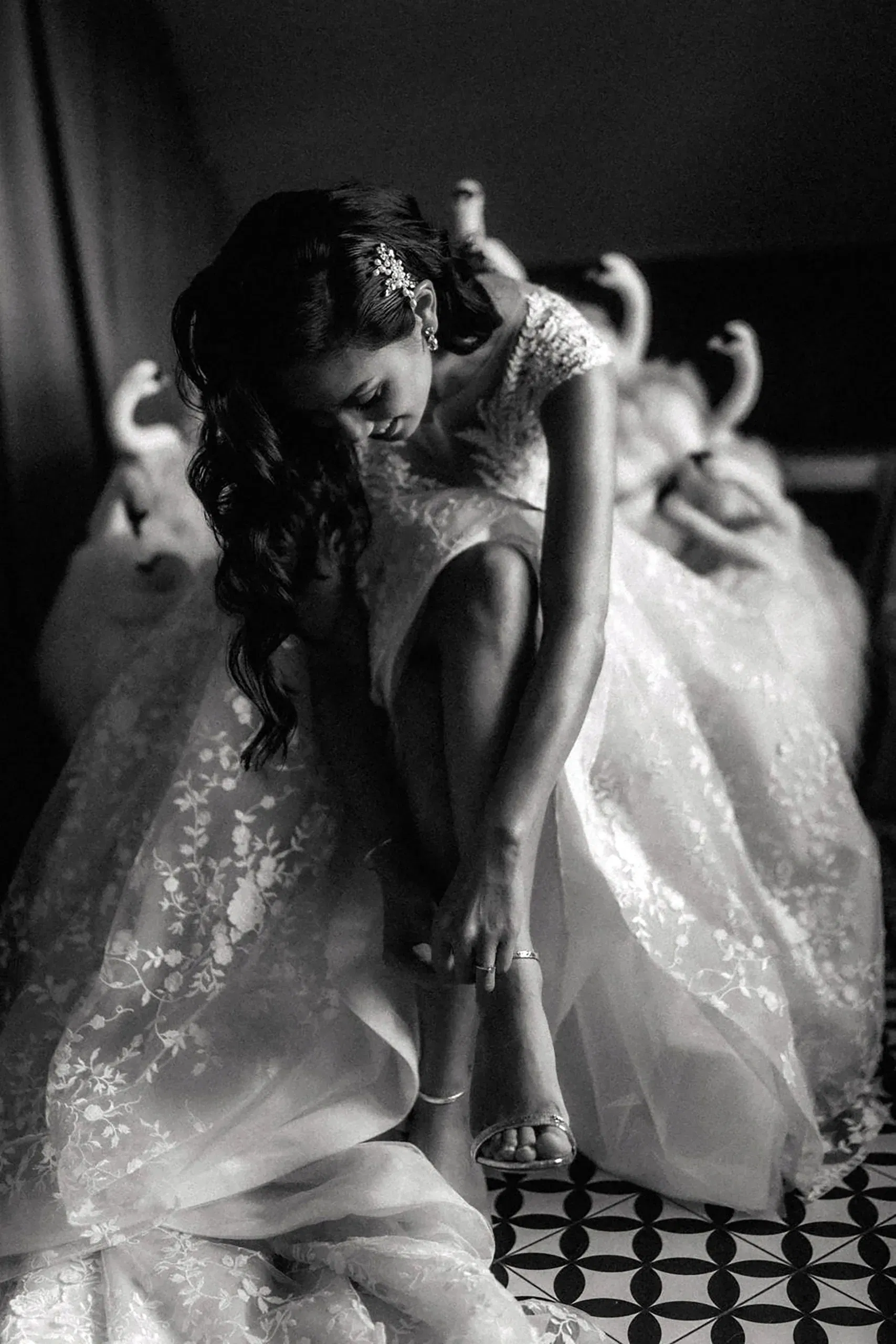 Schwarz-weiß-Aufnahme einer Braut, die sich im Sitzen ihre Sandalen schließt. Sie trägt ein besticktes, ausladendes Spitzen-Brautkleid und hat wellige Haare mit einem funkelnden Haaraccessoire zur Seite gesteckt.