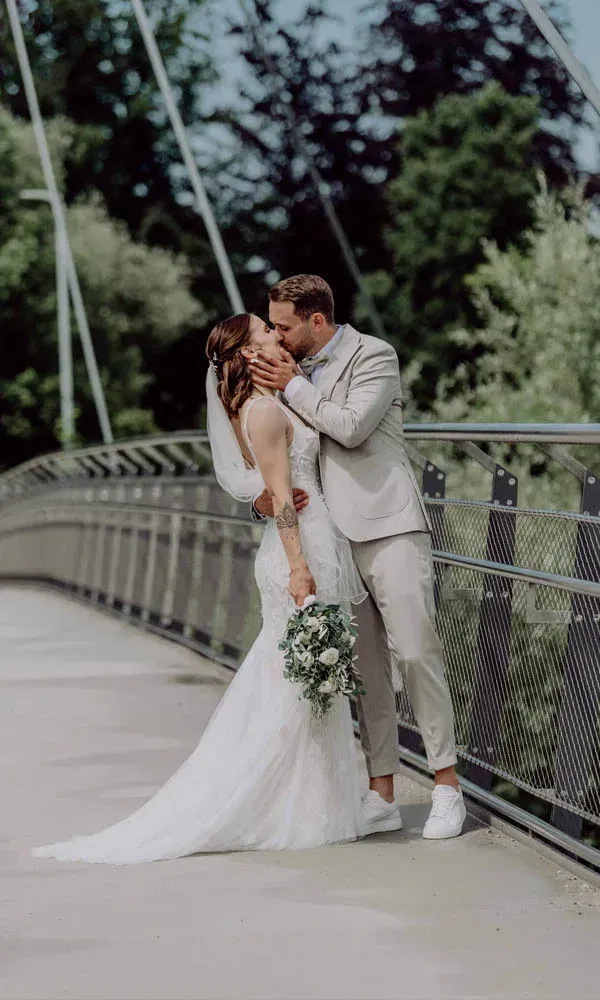 Braut und Bräutigam küssen sich innig auf einer modernen Brücke, umgeben von grüner Natur.