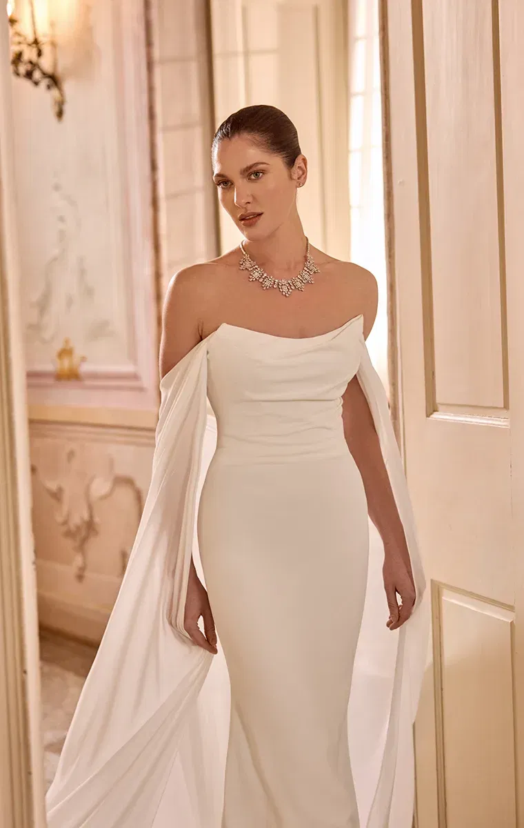 Modernes, schulterfreies Brautkleid mit glatter Silhouette und geschwungenen, fließenden Ärmeldrapierungen.