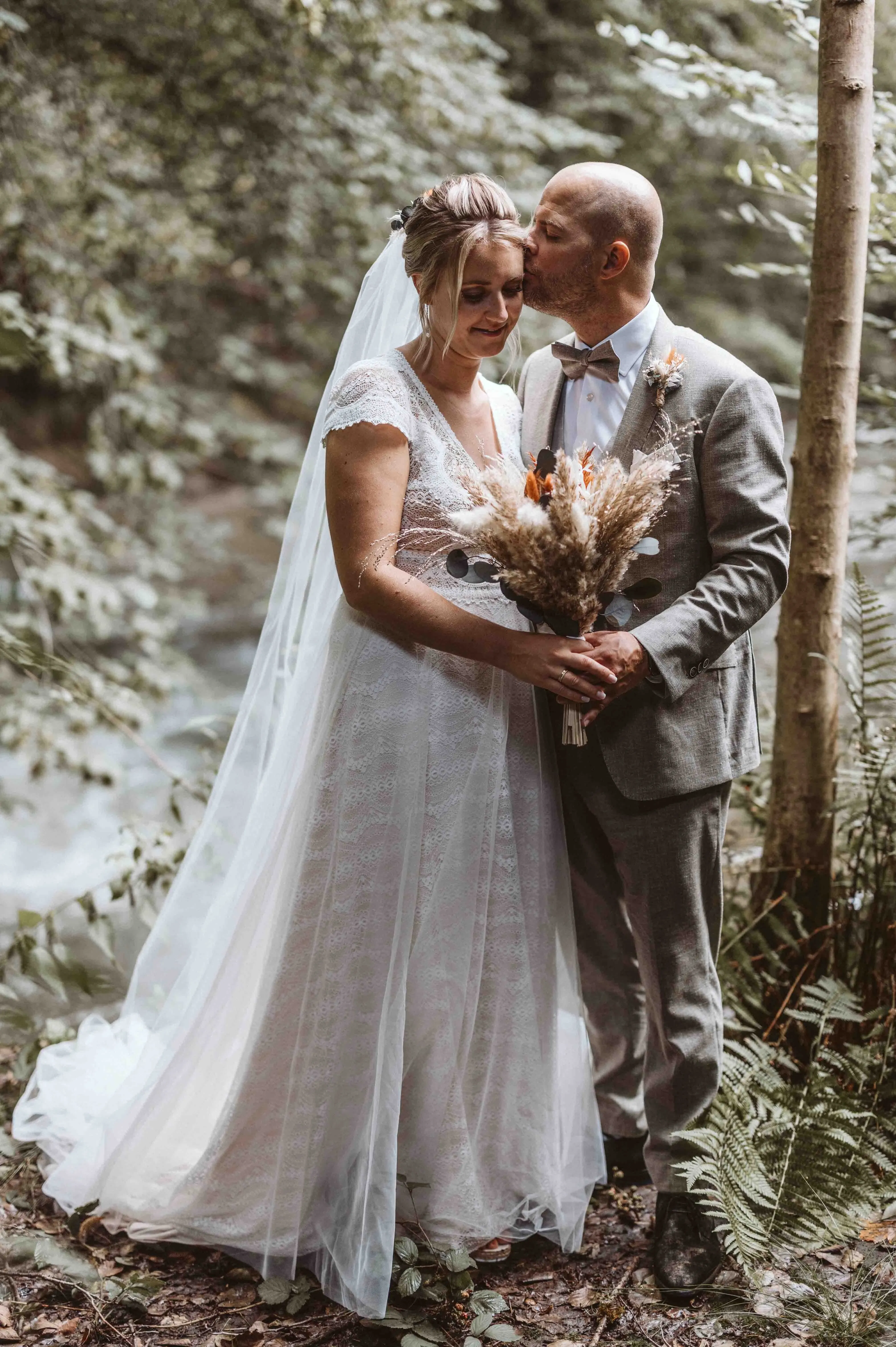 Braut und Bräutigam stehen inmitten eines grünen Waldstücks, während der Bräutigam der Braut sanft einen Kuss auf die Stirn gibt und sie den Trockenblumenstrauß hält.