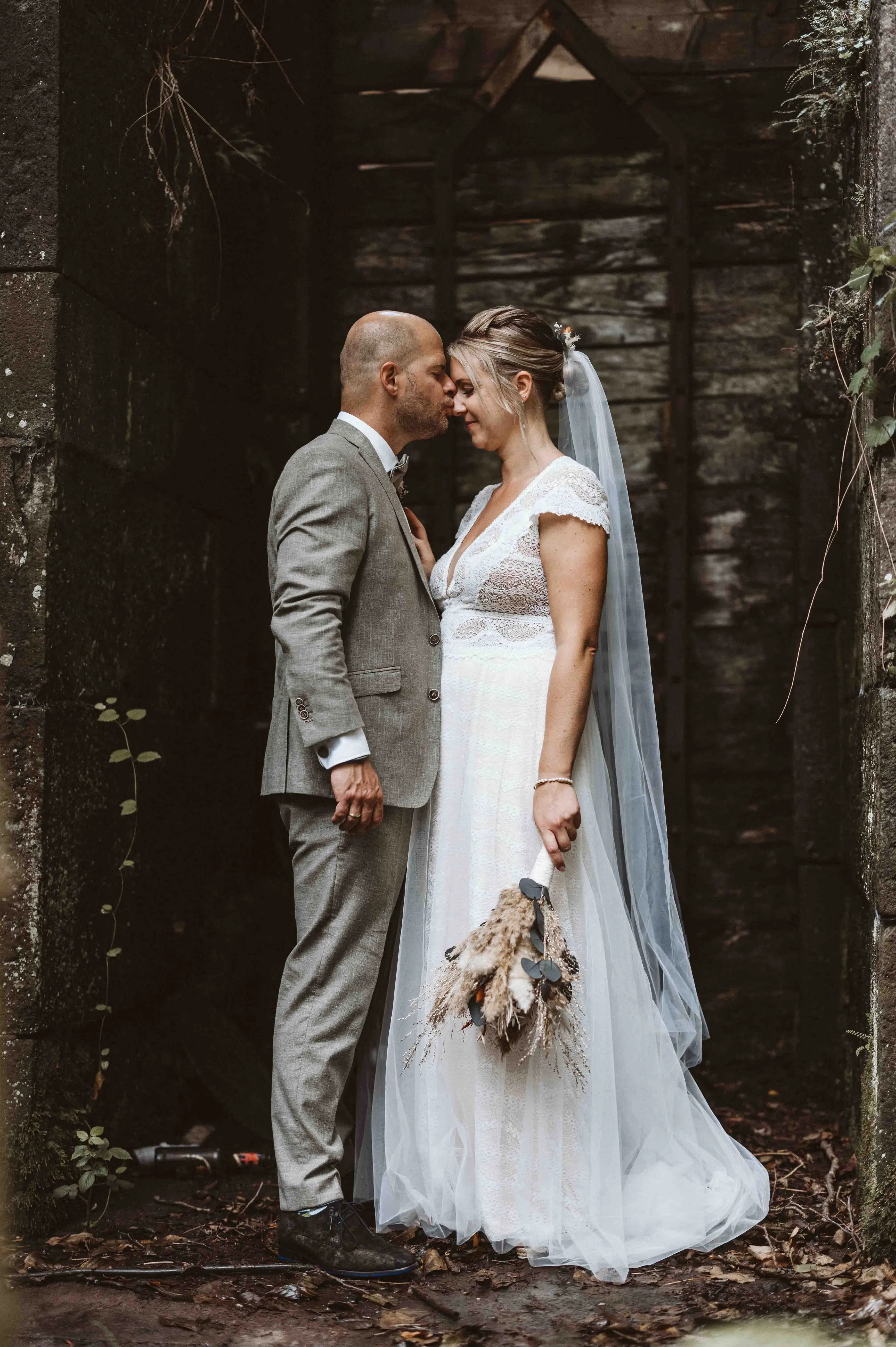 Braut und Bräutigam stehen nah beieinander in einer alten, moosbewachsenen Ruine, während er ihr sanft einen Kuss auf die Stirn gibt.