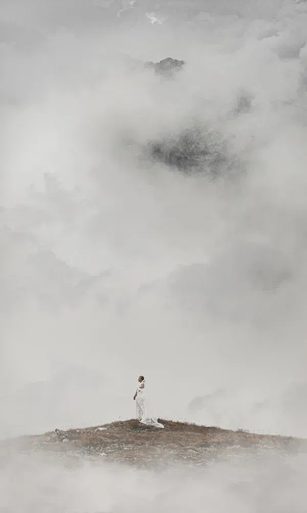 Braut in einem langen weißen Kleid steht allein auf einem nebelverhangenen Berggipfel, umgeben von dichter, mystischer Wolkenlandschaft.