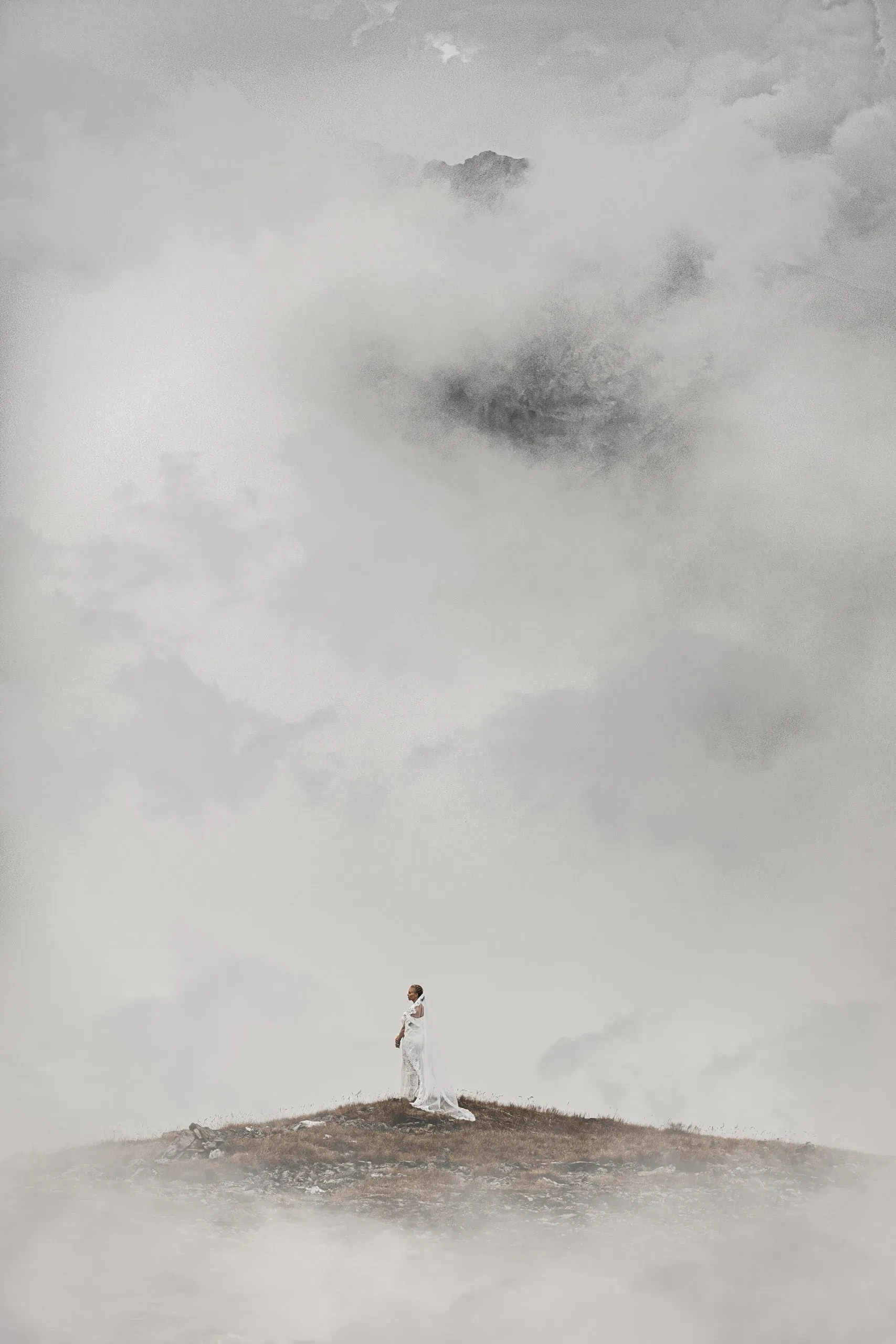 Braut in einem langen weißen Kleid steht allein auf einem nebelverhangenen Berggipfel, umgeben von dichter, mystischer Wolkenlandschaft.