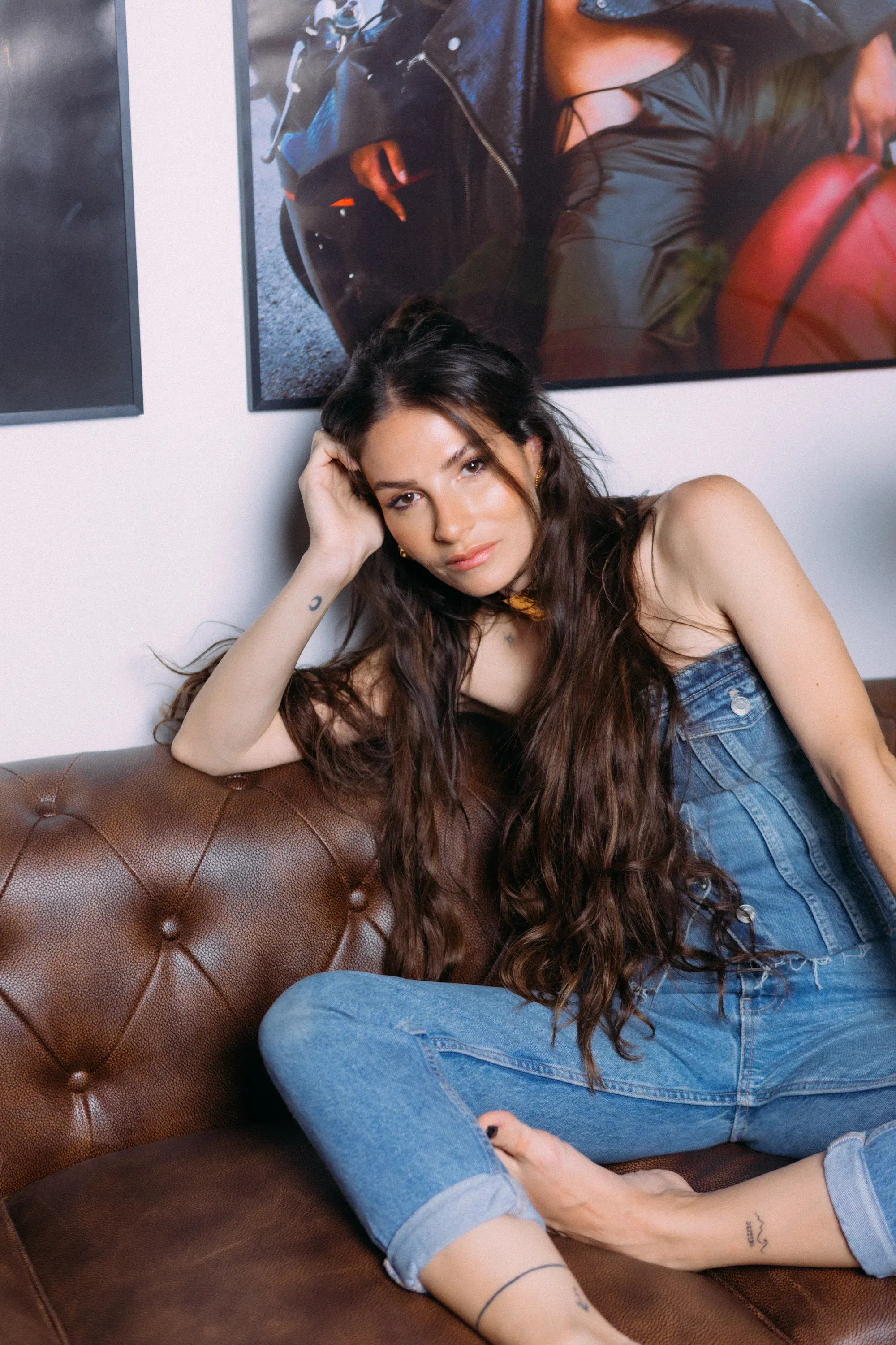 Frau in Jeans-Latzhose sitzt entspannt auf einem braunen Leder-Sofa und schaut direkt in die Kamera.