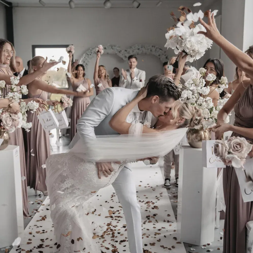 Bräutigam küsst seine Braut leidenschaftlich beim Auszug, während Freunde jubelnd Blütenblätter werfen.