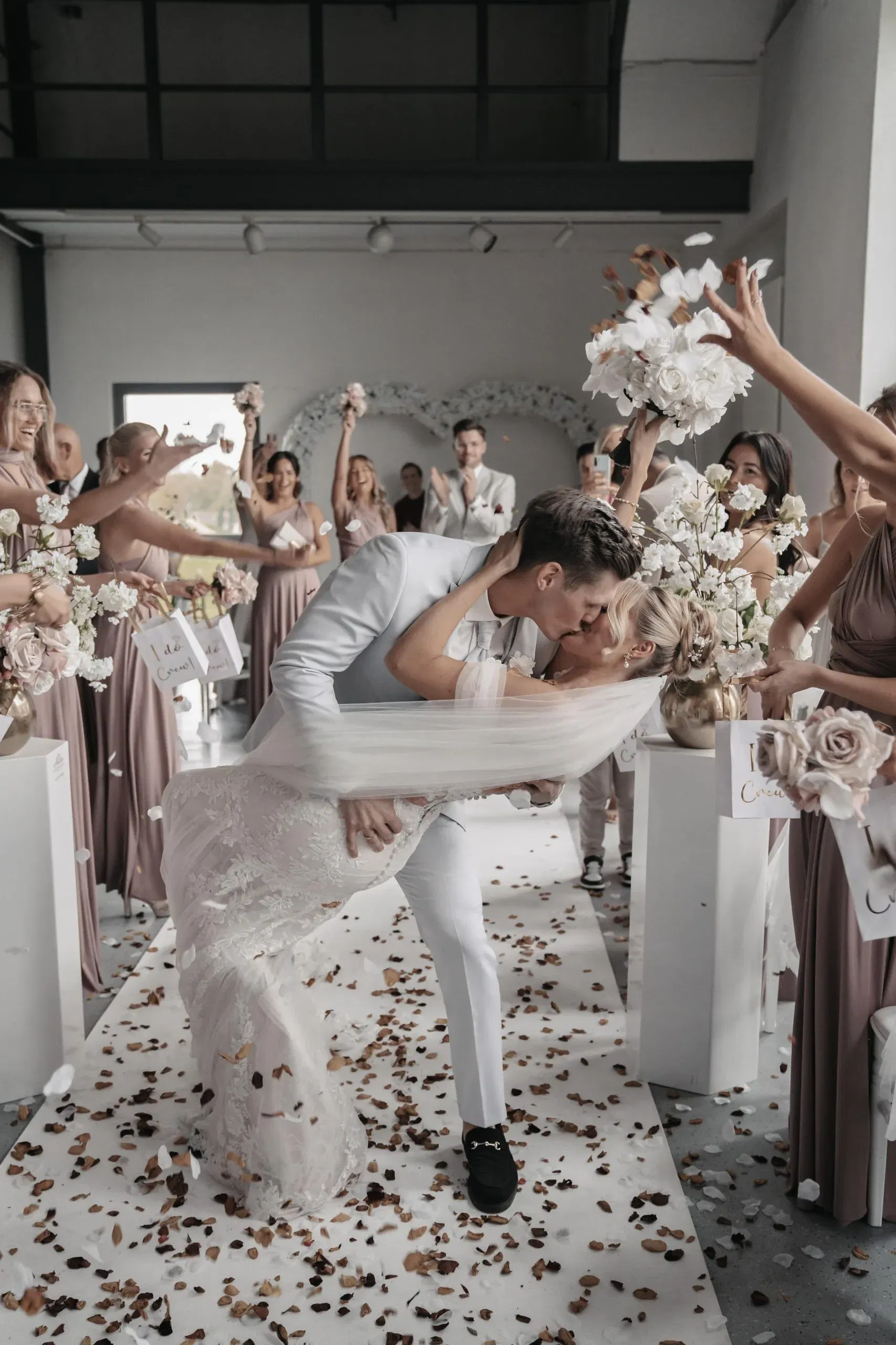 Bräutigam küsst seine Braut leidenschaftlich beim Auszug, während Freunde jubelnd Blütenblätter werfen.