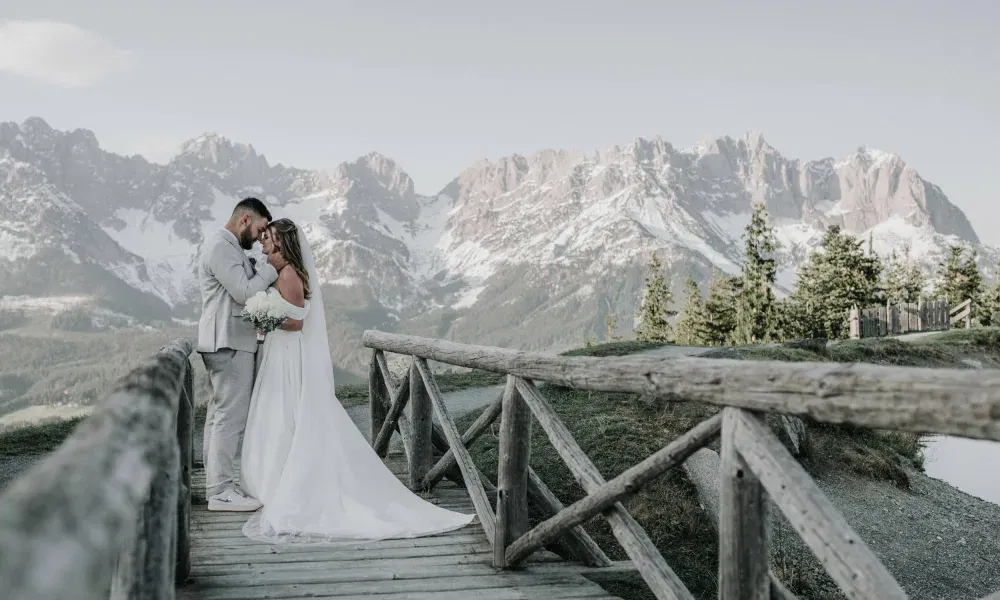 Braut und Bräutigam stehen eng umschlungen auf einer Holzbrücke vor einer eindrucksvollen Bergkulisse mit schneebedeckten Gipfeln und halten sich liebevoll an den Händen.