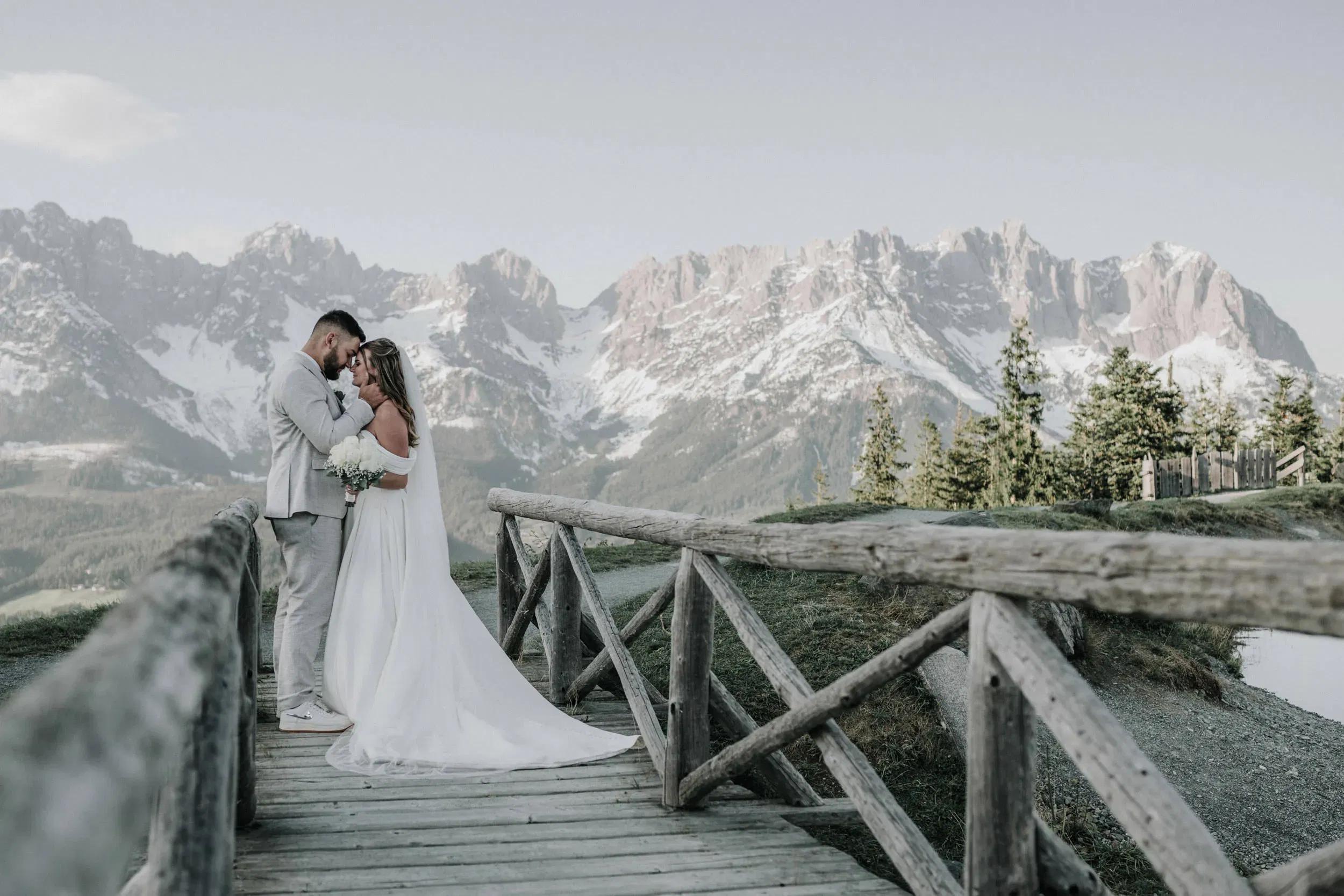 Braut und Bräutigam stehen eng umschlungen auf einer Holzbrücke vor einer eindrucksvollen Bergkulisse mit schneebedeckten Gipfeln und halten sich liebevoll an den Händen.