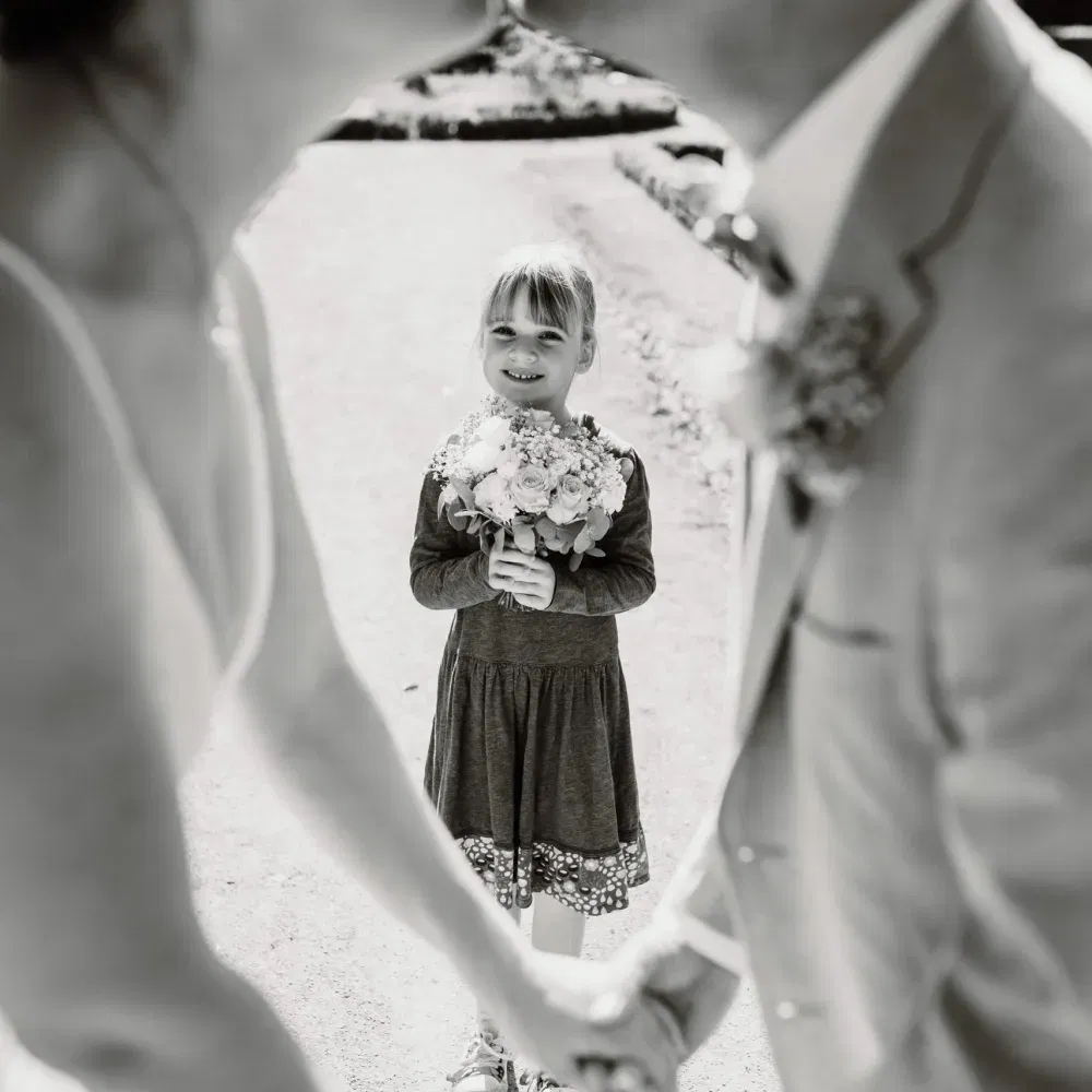 Schwarz-Weiß-Foto eines Brautpaars, das sich küsst, während ein lächelndes Kind mit Blumenstrauß zwischen ihnen steht.