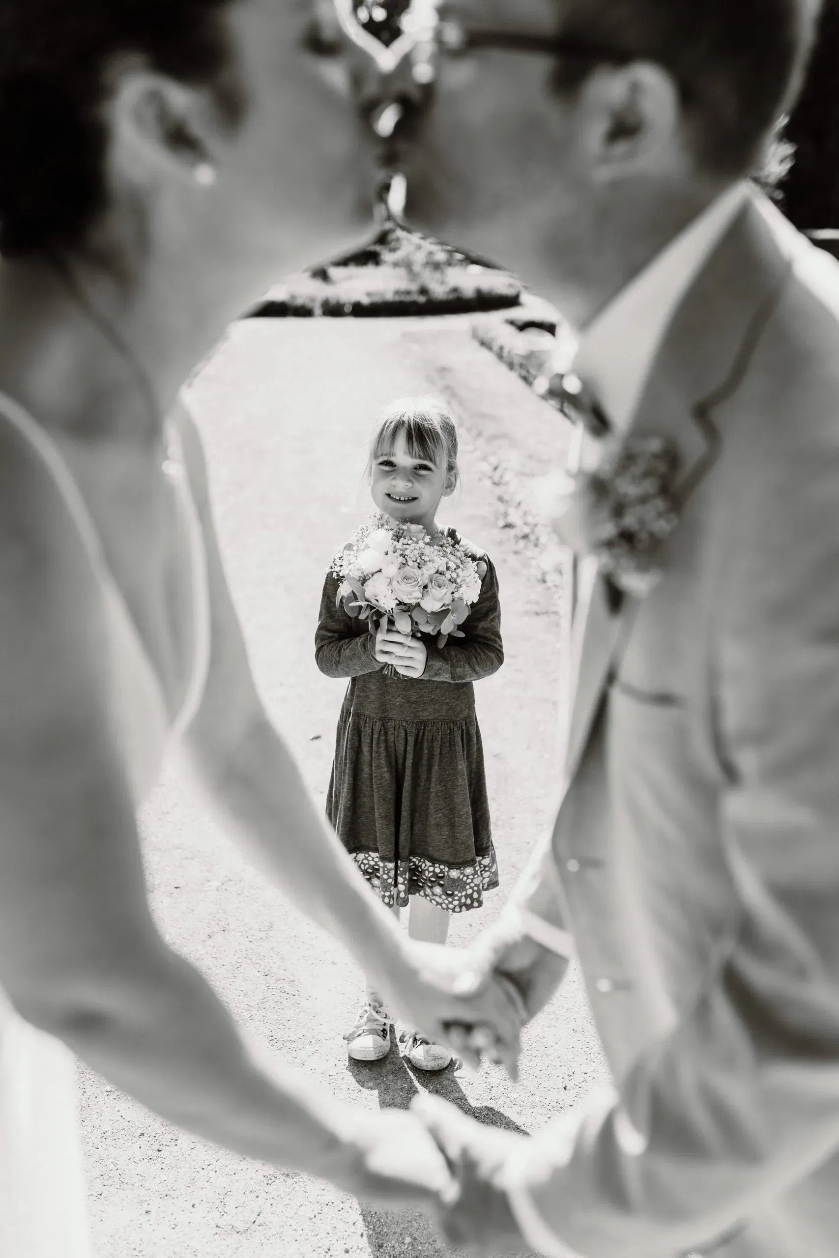 Schwarz-Weiß-Foto eines Brautpaars, das sich küsst, während ein lächelndes Kind mit Blumenstrauß zwischen ihnen steht.