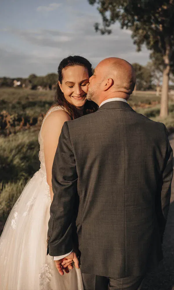 Braut und Bräutigam stehen im warmen Abendlicht auf einem Weg, halten Händchen und der Bräutigam küsst die Braut zärtlich auf die Wange.