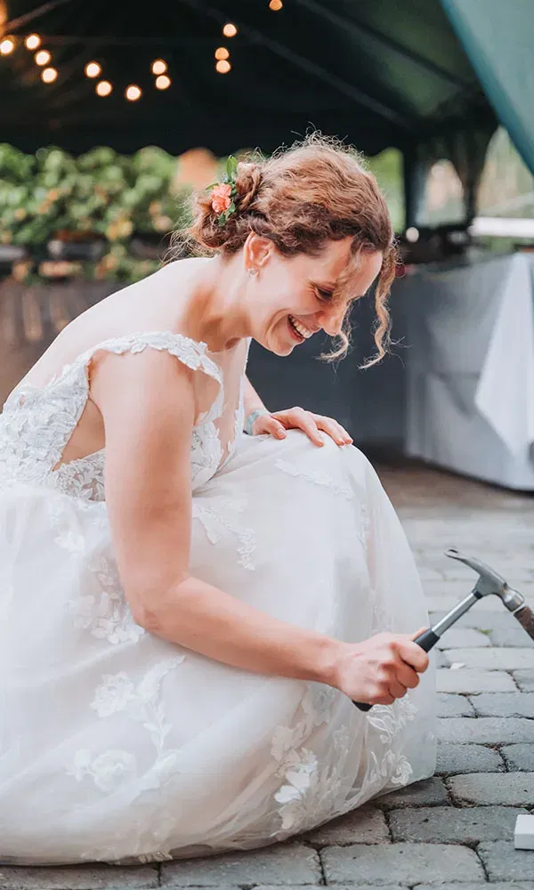 Die Braut sitzt lachend im Hochzeitskleid am Boden und hält einen Hammer in der Hand.