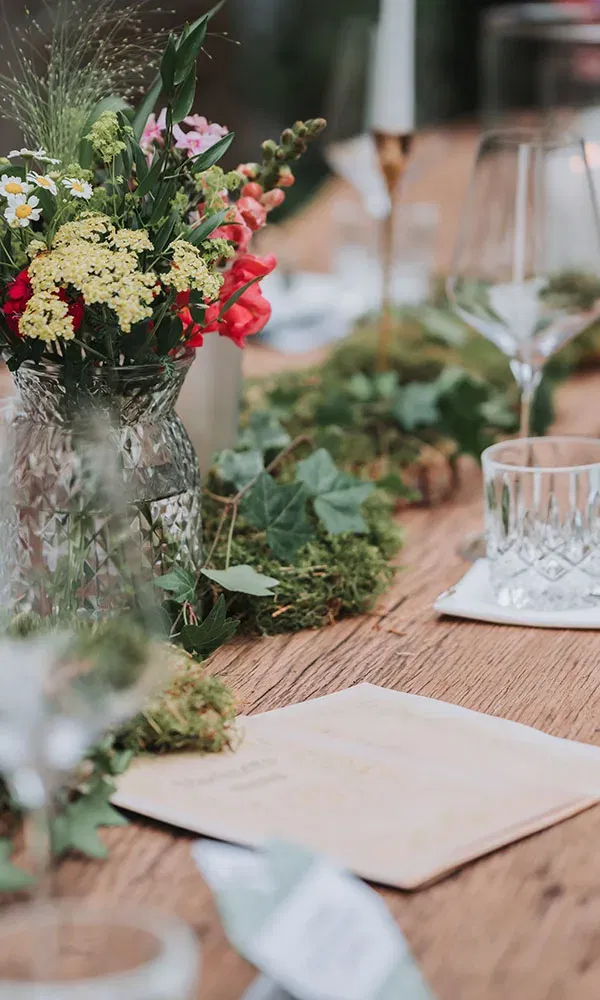 Festlich gedeckter Holztisch mit einer Vase voller bunter Blumen und natürlicher Deko aus Moos und Efeu.