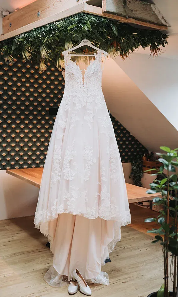 Elegantes Hochzeitskleid mit floralen Details auf einem Bügel, darunter die weißen Schuhe der Braut.