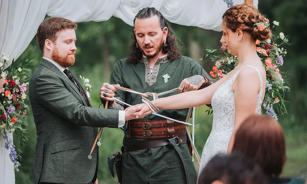 Braut und Bräutigam nehmen an einer Handfasting-Zeremonie teil, während der Trauredner bunte Bänder um ihre Hände legt.