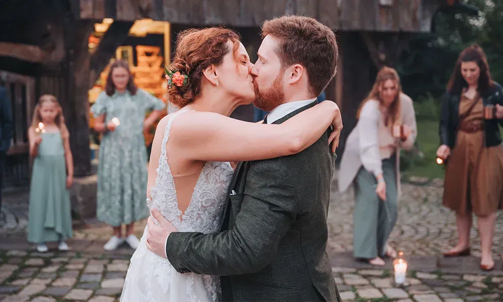 Braut und Bräutigam küssen sich innig bei der Abendfeier, umgeben von Gästen mit kleinen Lichtern.