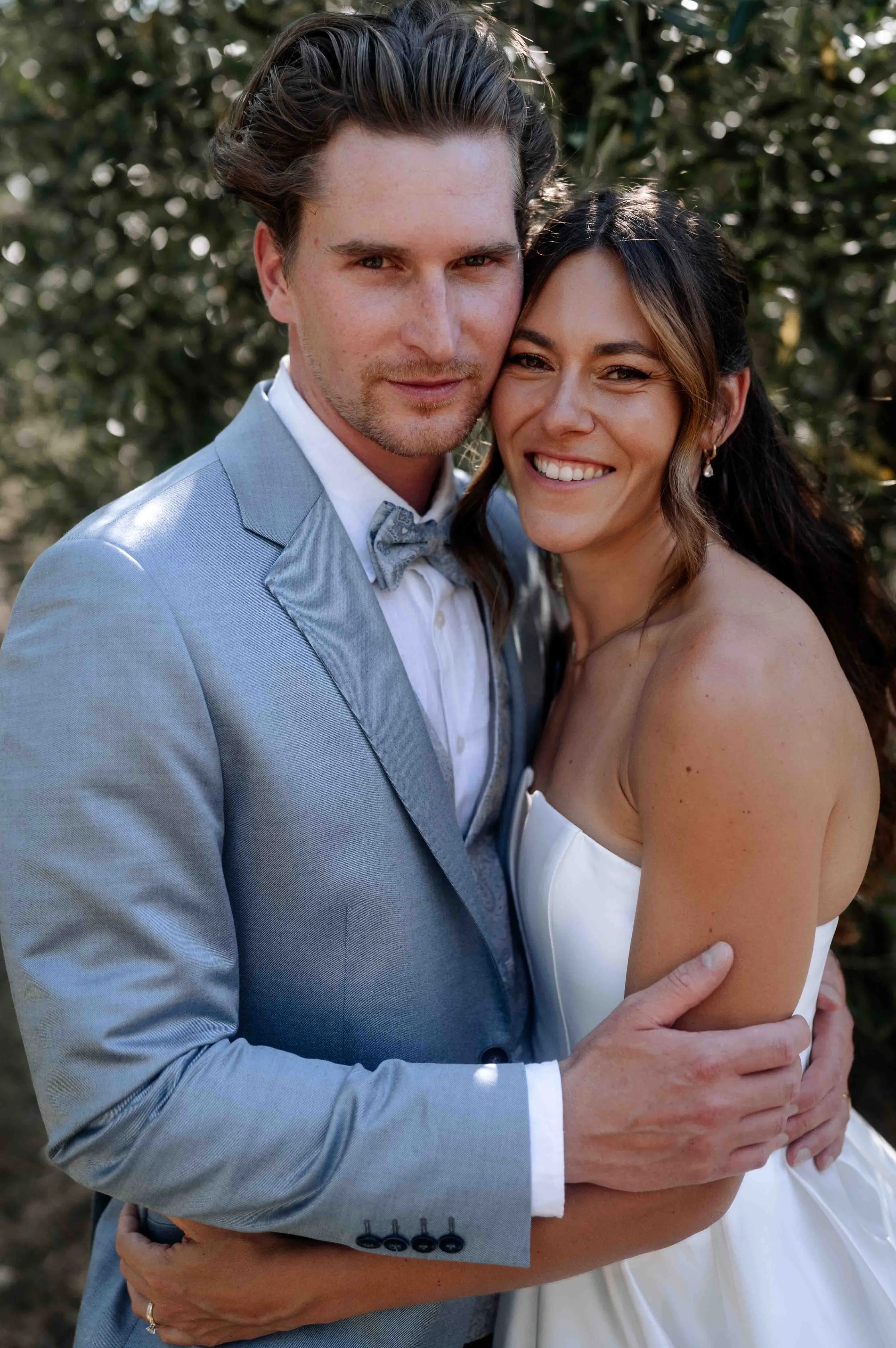 Braut und Bräutigam umarmen sich eng und lächeln in die Kamera, umgeben von weichem Naturlicht und grünen Blättern im Hintergrund.