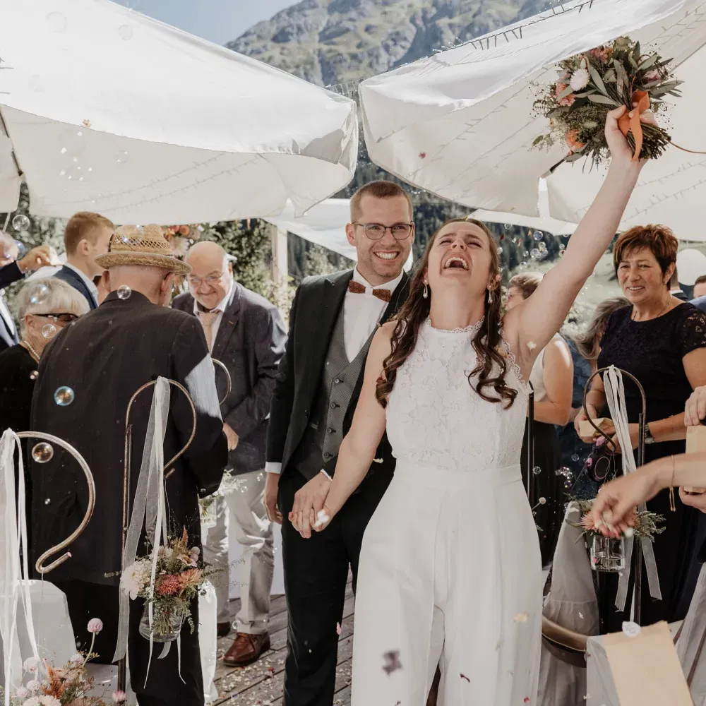 Frisch vermähltes Paar geht lachend durch ein Spalier aus Gästen; die Braut hebt jubelnd ihren Brautstrauß in die Luft, während Seifenblasen und Blütenkonfetti durch die Sonne schweben.