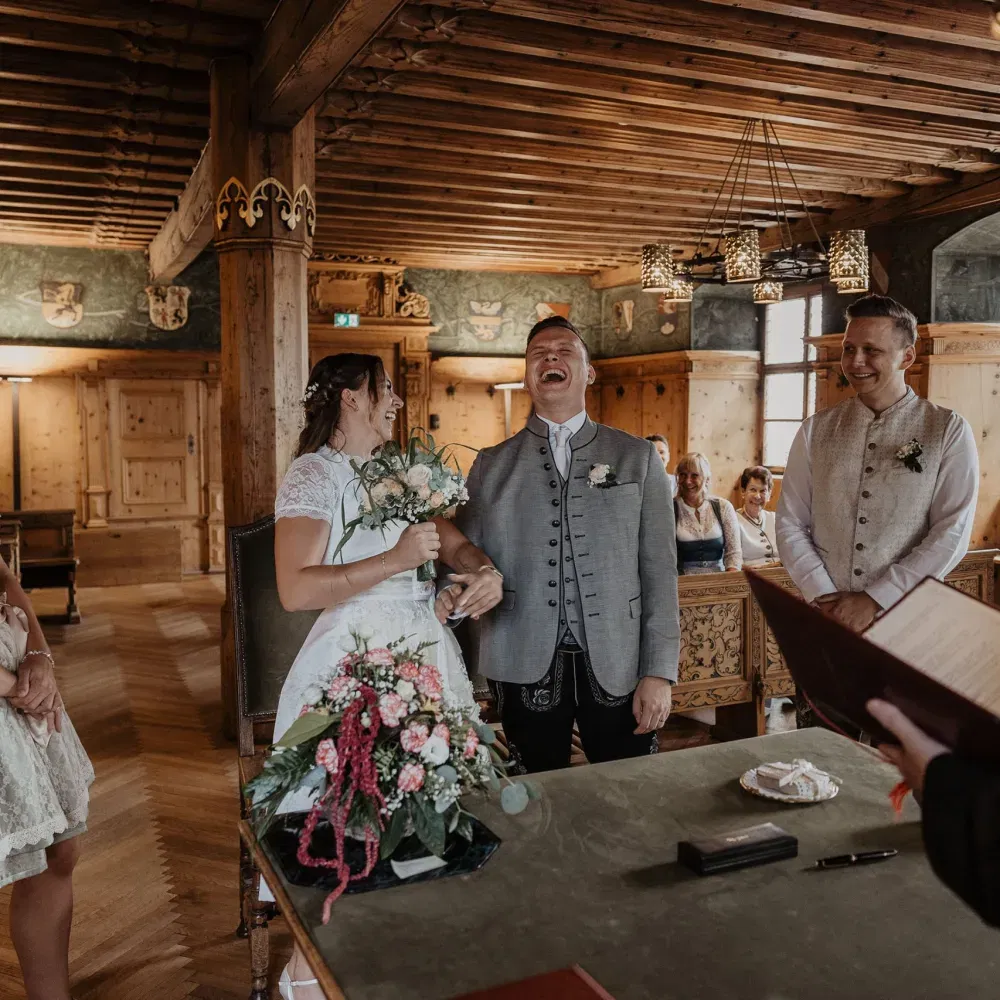 Fröhliches Brautpaar in Tracht lacht während der standesamtlichen Trauung in einem holzgetäfelten Raum.