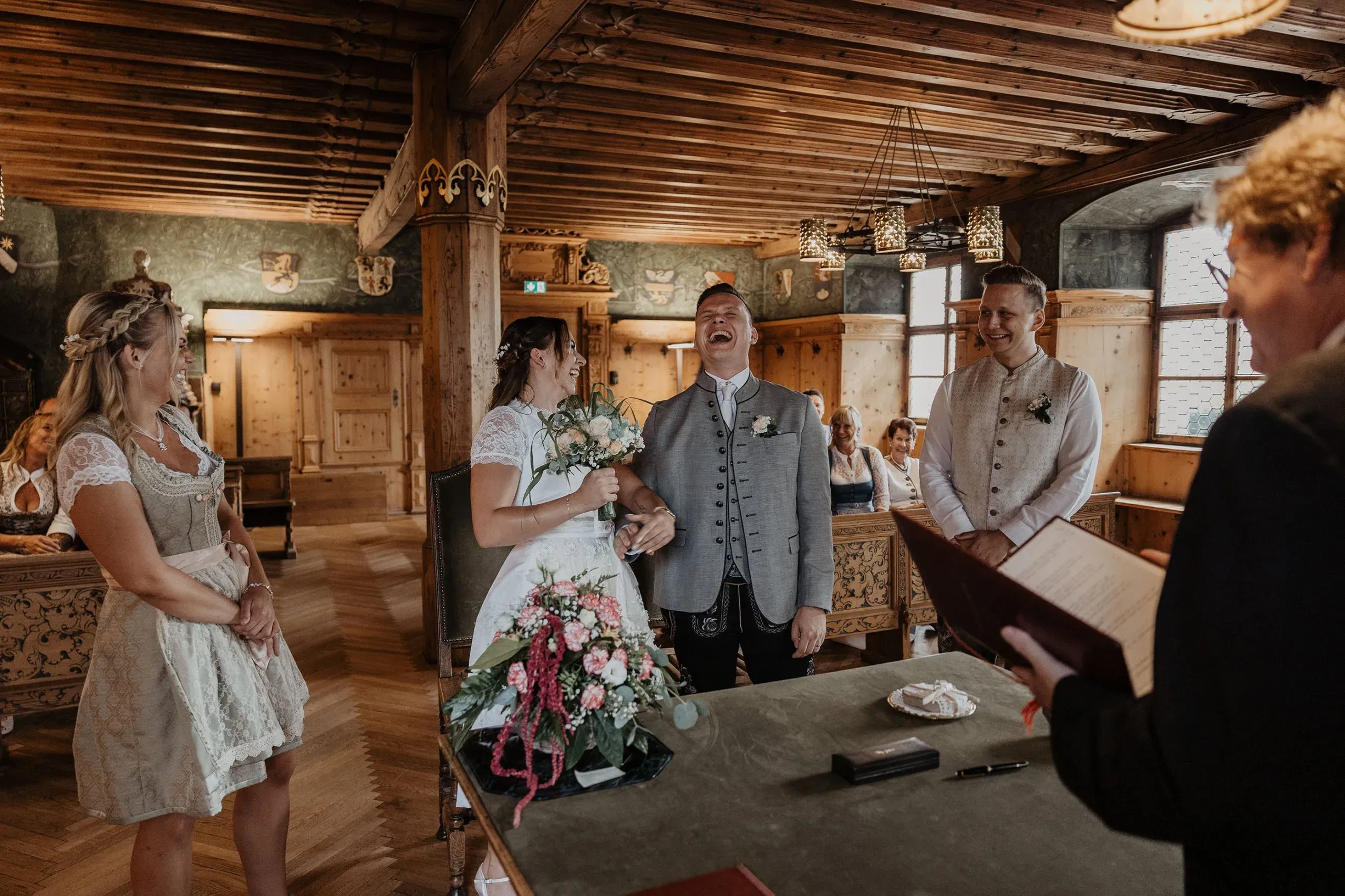 Fröhliches Brautpaar in Tracht lacht während der standesamtlichen Trauung in einem holzgetäfelten Raum.