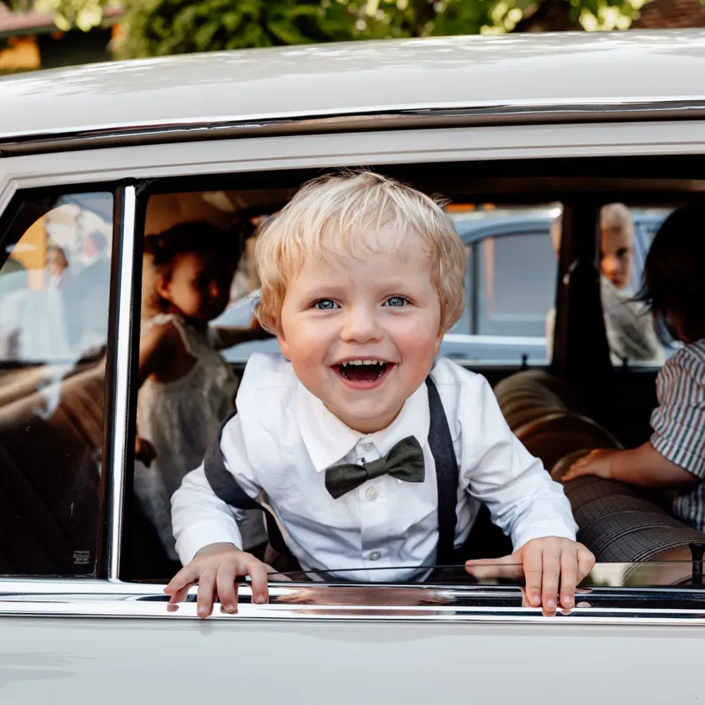 Fröhlicher Junge im Hemd und Fliege lacht aus dem Fenster eines Oldtimers bei einer Hochzeit.