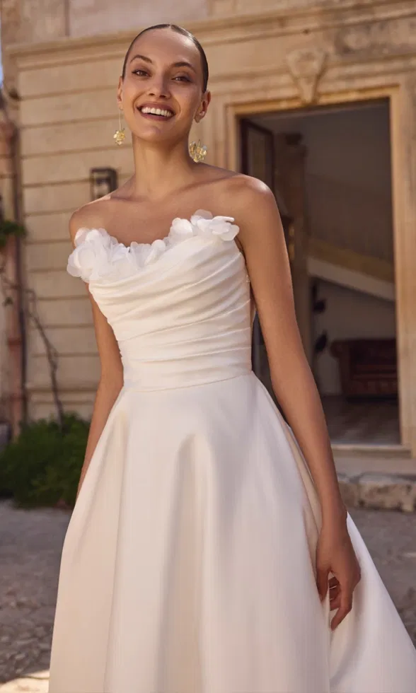 Brautmodell trägt ein modernes, trägerloses Brautkleid mit weich drapierter Corsage und floralen 3D-Applikationen am Ausschnitt, kombiniert mit fließendem Rock, fotografiert vor einer mediterranen Steinarchitektur im warmen Tageslicht.