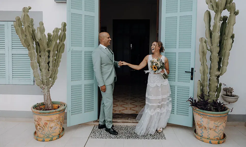 Ein Hochzeitspaar vor türkisfarbenen Fensterläden: Die Braut im luftigen Spitzenkleid greift nach der Hand des Bräutigams im hellgrünen Anzug und lächelt ihn herzlich an.