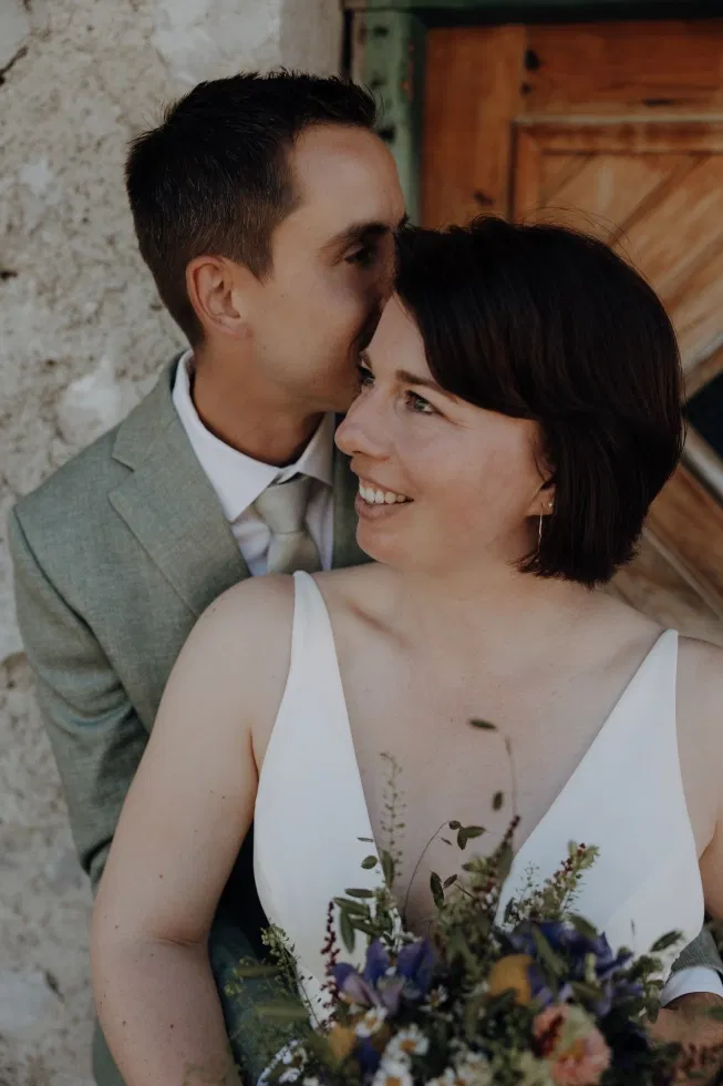 Zärtliches Porträt eines Brautpaares, bei dem der Bräutigam der Braut liebevoll einen Kuss auf die Stirn gibt, während sie lächelnd in die Ferne blickt.