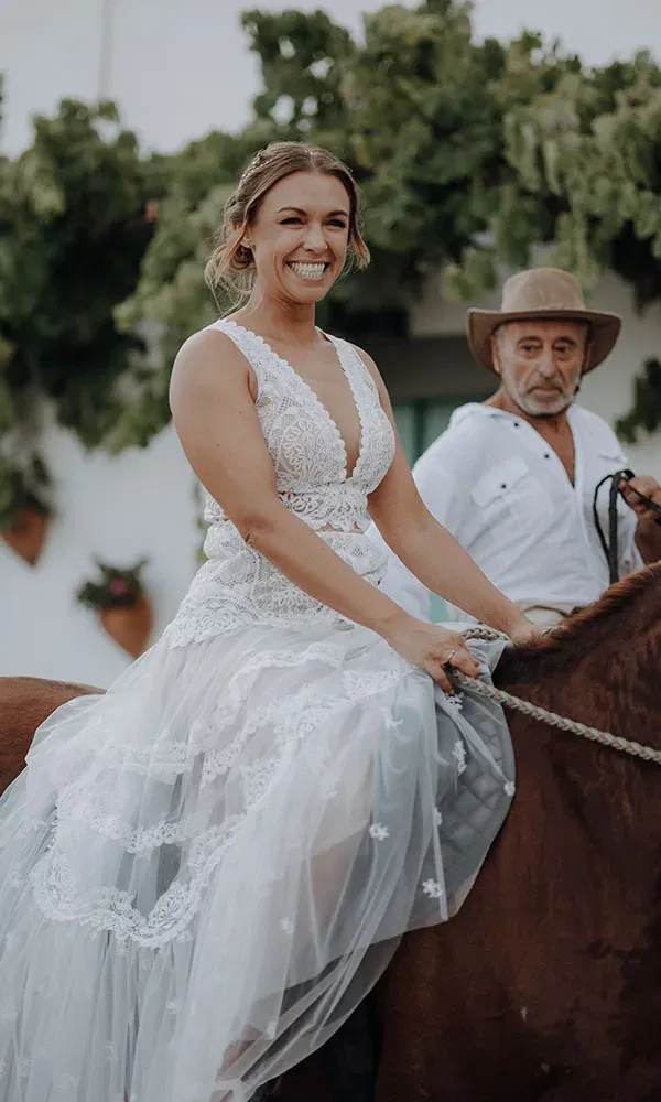Die Braut sitzt strahlend im Spitzenkleid auf einem Pferd, während ein Reitführer neben ihr reitet.