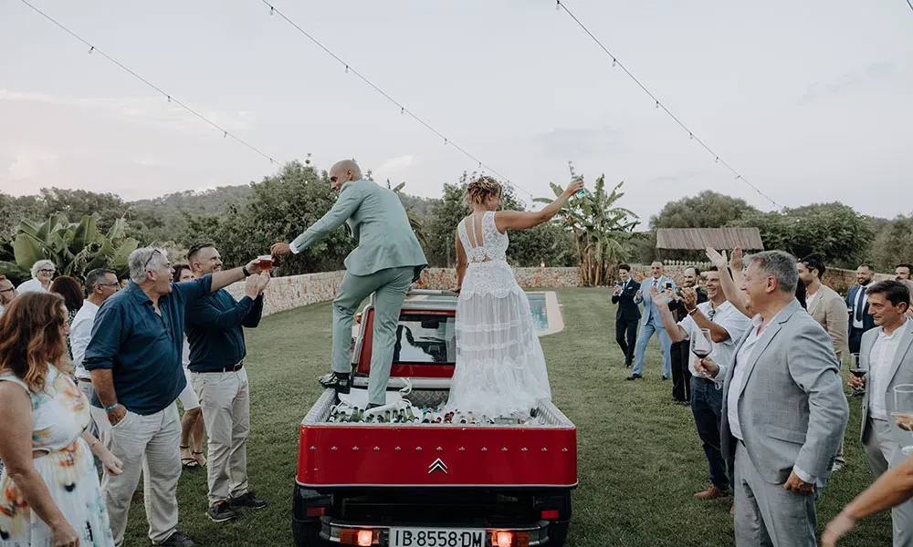 Hochzeitspaar feiert auf dem Rücken eines roten Autos, umgeben von jubelnden Gästen, die ihre Gläser in die Höhe halten.