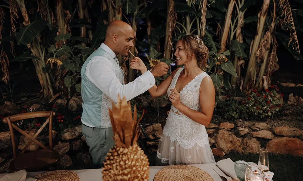 Braut und Bräutigam teilen einen süßen Bissen und lachen, eingerahmt von üppigem Grün und romantischer Abendstimmung.