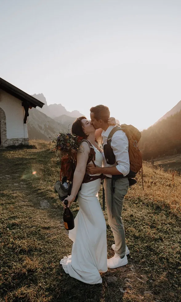 Braut und Bräutigam küssen sich mit Wanderrucksäcken auf dem Rücken, während die Sonne hinter den Bergen untergeht.