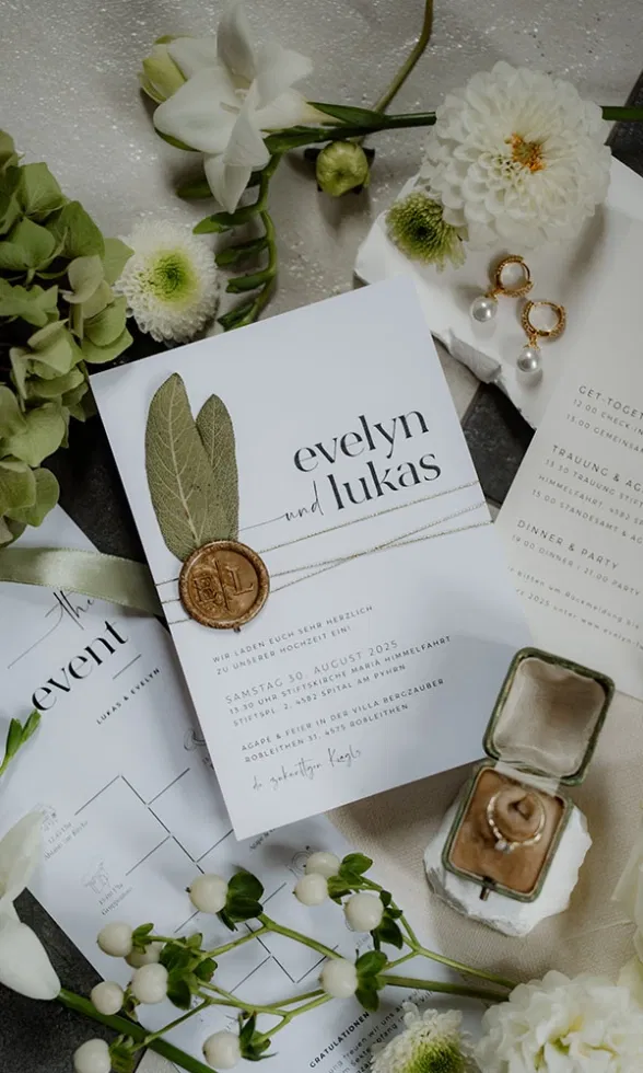 Flatlay der Hochzeitspapeterie mit Einladung „Evelyn und Lukas“, Wachssiegel, floralen Details und Eheringen.