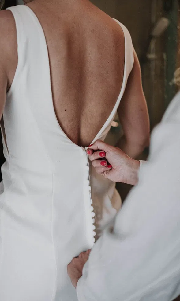 Detailaufnahme der Rückseite eines schlichten Brautkleides mit tiefem Rückenausschnitt, während eine helfende Hand die feinen Knöpfe schließt.