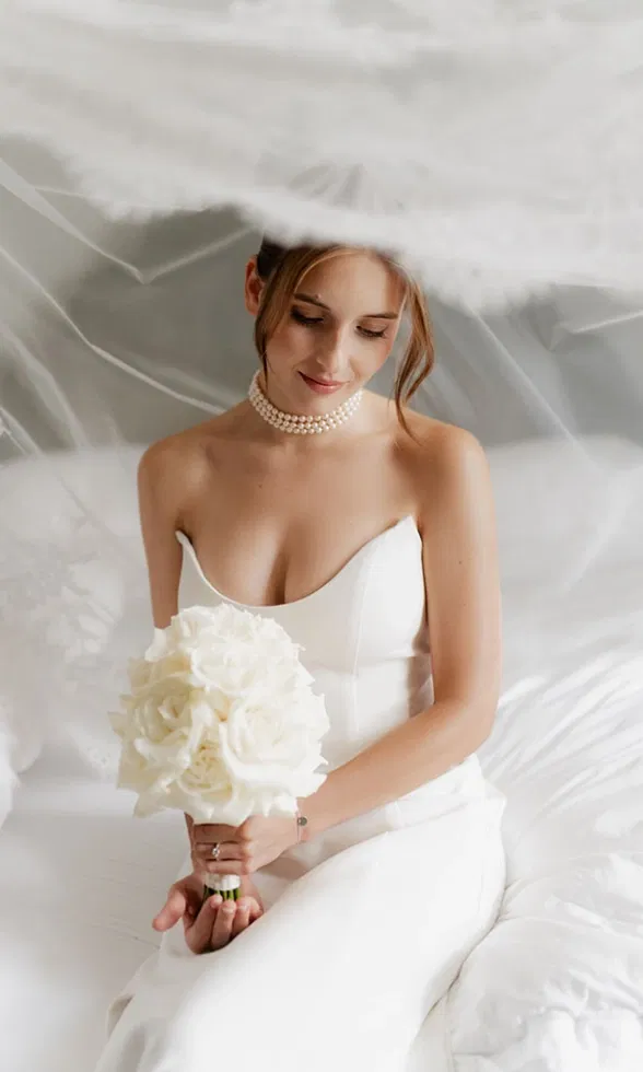 Sanftes Brautporträt mit Schleier: Die Braut sitzt im schlichten Brautkleid und hält einen weißen Brautstrauß in ruhiger, eleganter Atmosphäre.