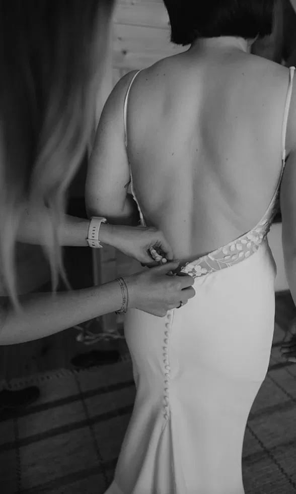 Eine Person hilft der Braut beim Schließen der Knöpfe am Rücken ihres eleganten Kleides.
