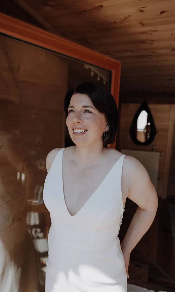 Die Braut steht lächelnd im warmen Licht einer Holzhütte und trägt ihr schlichtes Kleid mit tiefem Ausschnitt.