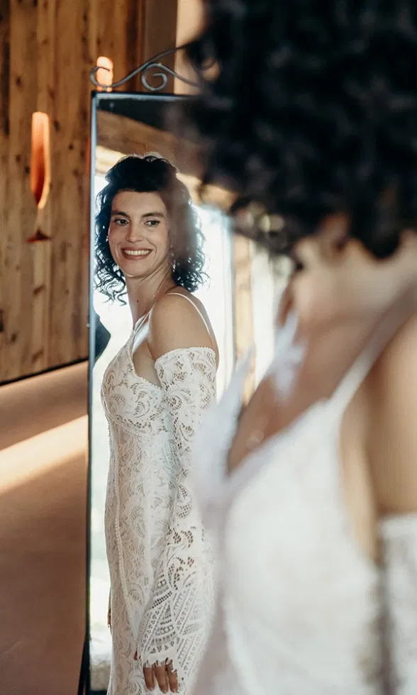 Braut im Spitzenbrautkleid betrachtet sich lächelnd im Spiegel während des Getting Ready, warmer Lichteinfall sorgt für eine intime Atmosphäre.