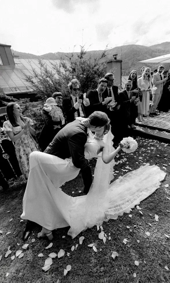 Schwarz-weiß Hochzeitsmoment: Das Brautpaar küsst sich ausgelassen, während die Gäste jubeln und fotografieren.