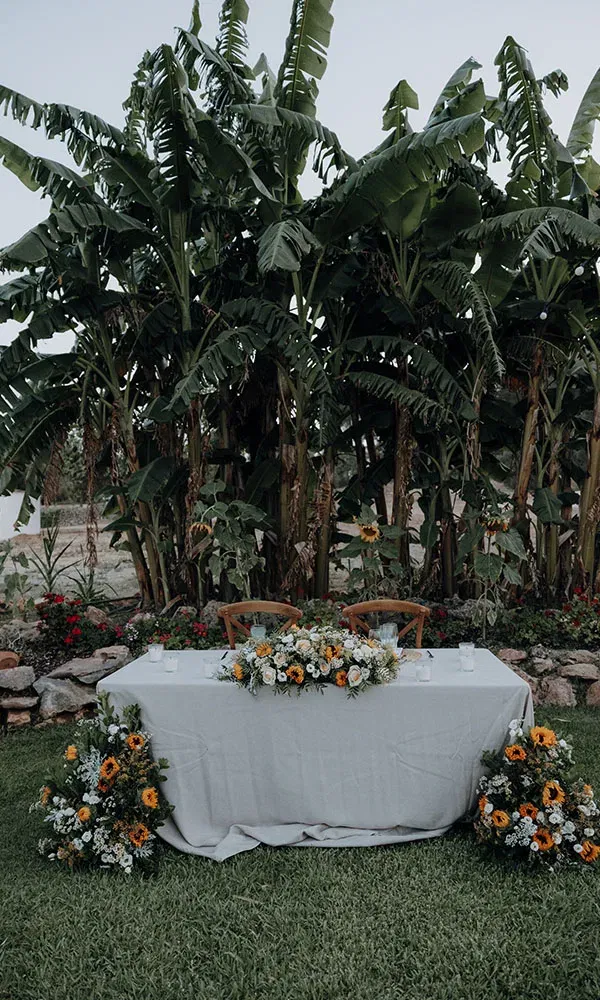 Geschmückter Hochzeitstisch im Garten mit üppigen Sonnenblumen-Arrangements vor einer Kulisse aus hohen Bananenstauden.
