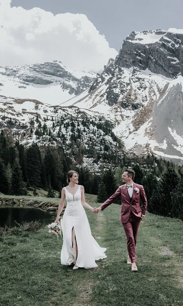 Braut und Bräutigam gehen Hand in Hand durch eine alpine Wiesenlandschaft, im Hintergrund ein Bergsee, Tannen und schneebedeckte Gipfel.