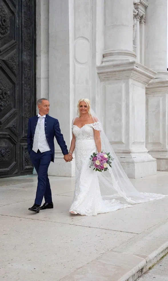 Braut und Bräutigam gehen Hand in Hand vor der imposanten Fassade einer venezianischen Kirche, die Braut trägt ein elegantes Spitzenbrautkleid mit Schleier und Blumenstrauß, der Bräutigam einen dunkelblauen Anzug – ein zeitloser Hochzeitsmoment vor historischer Kulisse.