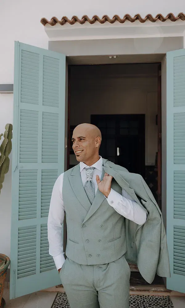 Bräutigam steht vor mintfarbenen Türen, trägt seine Anzugjacke lässig über der Schulter und lächelt entspannt zur Seite.