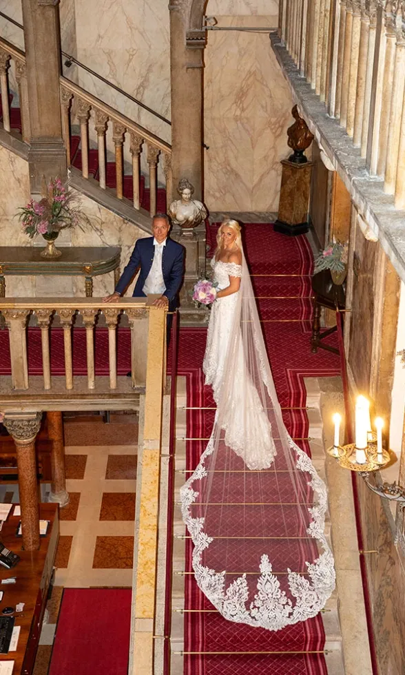 Braut und Bräutigam posieren auf einer prunkvollen Treppe im Inneren einer historischen Kirche, die Braut im Spitzenbrautkleid mit langem Schleier und ausladender Schleppe, der Bräutigam im dunkelblauen Anzug – ein edler Hochzeitsmoment in luxuriösem Ambiente.