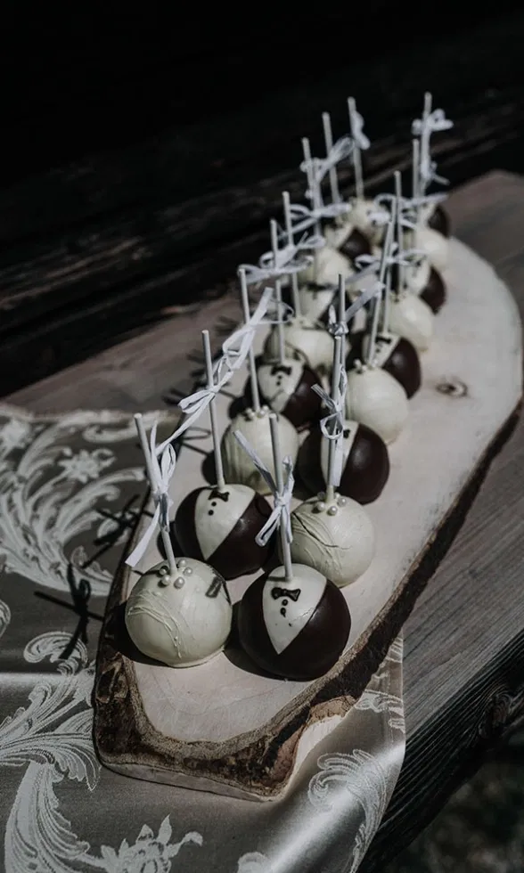Kreative Hochzeitsdesserts in Form von Cake Pops, dekoriert als Braut und Bräutigam, arrangiert auf einem rustikalen Holzbrett.