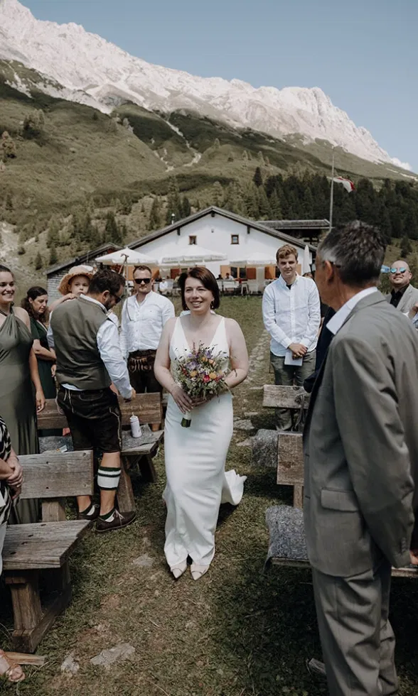 Die Braut schreitet mit Blumenstrauß zwischen den Gästen über eine Wiese, im Hintergrund eine Berghütte und hohe Gipfel.