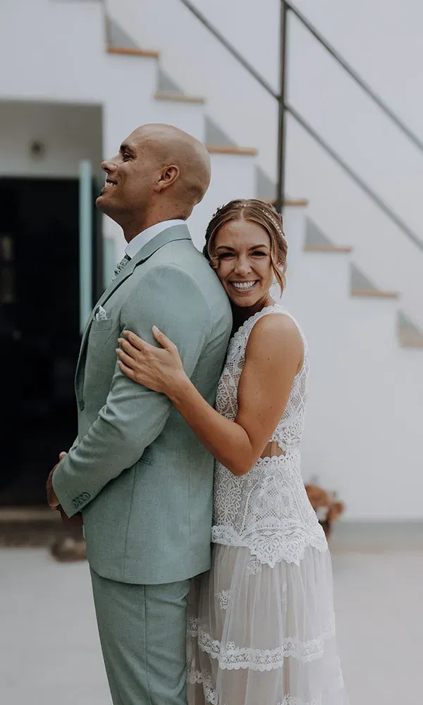 Braut und Bräutigam stehen Rücken an Rücken, die Braut umarmt ihn strahlend und lehnt ihren Kopf an seine Schulter.