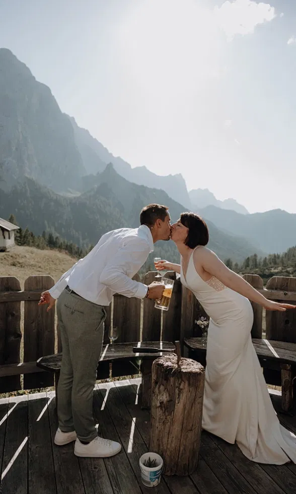 Braut und Bräutigam küssen sich auf einer Holzterrasse in den Bergen, während sie Bierkrüge in der Hand halten und die Sonne hinter den Gipfeln steht.